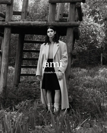 An adult female stands outside a rustic wooden tower, wearing an oversized coat, black skirt, turtleneck, and socks in black and white.