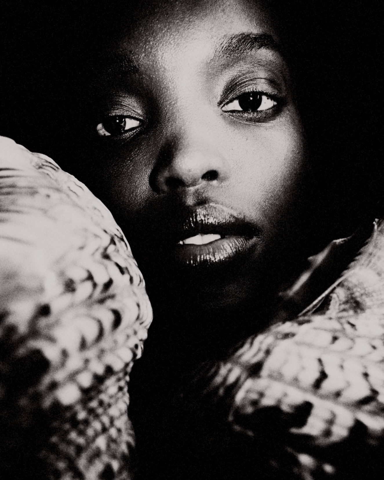 Close-up black-and-white portrait of a Black woman with dark eyes and glossy lips, framed by textured objects, creating a dramatic visual.