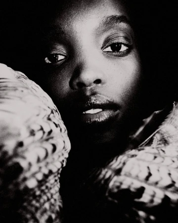 Close-up black-and-white portrait of a Black woman with dark eyes and glossy lips, framed by textured objects, creating a dramatic visual.