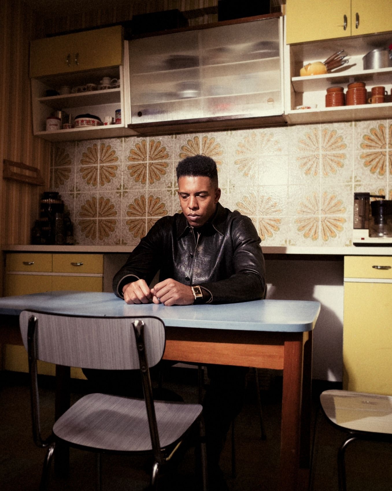 Man in a black leather jacket sits at a light blue table in a kitchen with tile walls and yellow cabinets, pensive mood, indoor setting.