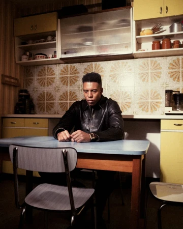 Man in a black leather jacket sits at a light blue table in a kitchen with tile walls and yellow cabinets, pensive mood, indoor setting.
