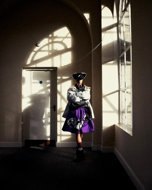 Woman stands in a naturally lit hallway wearing a hat, jacket, and a purple floral skirt with crossed arms next to a window and a door.