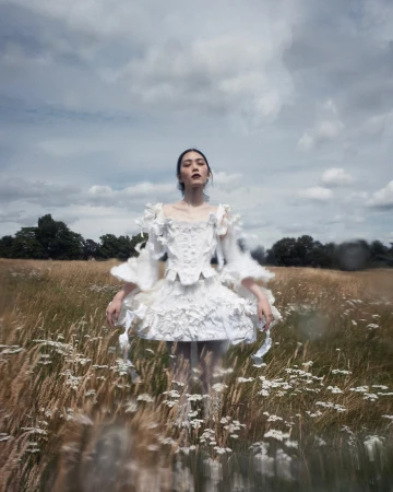 Woman in elaborate white dress stands in a field of wildflowers under a cloudy sky, creating a dreamy and ethereal aesthetic.