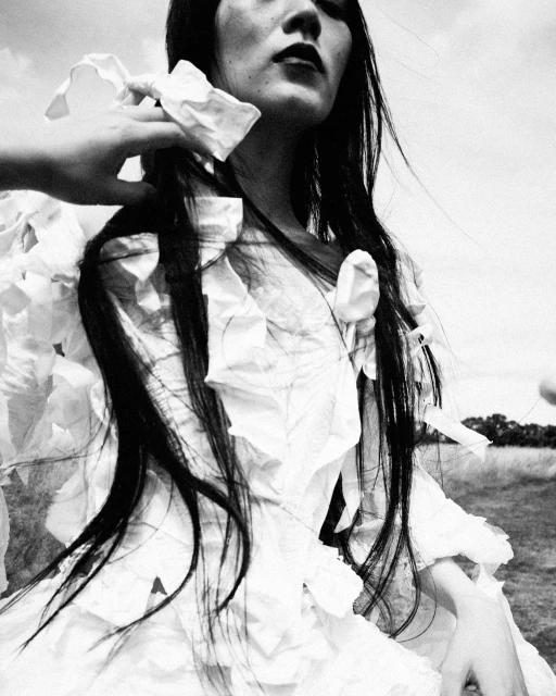 Black and white portrait of an Asian female model in a white dress, posing outdoors with a hand near her shoulder, emphasizing beauty and fashion.