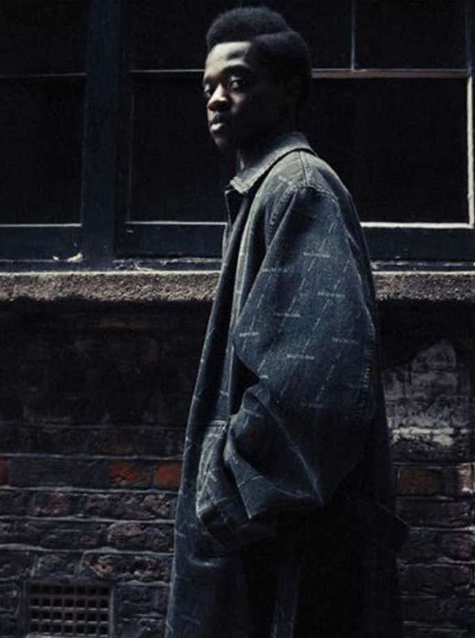 Young adult male poses in a printed overcoat in front of a brick wall and dark window, capturing urban fashion and style.