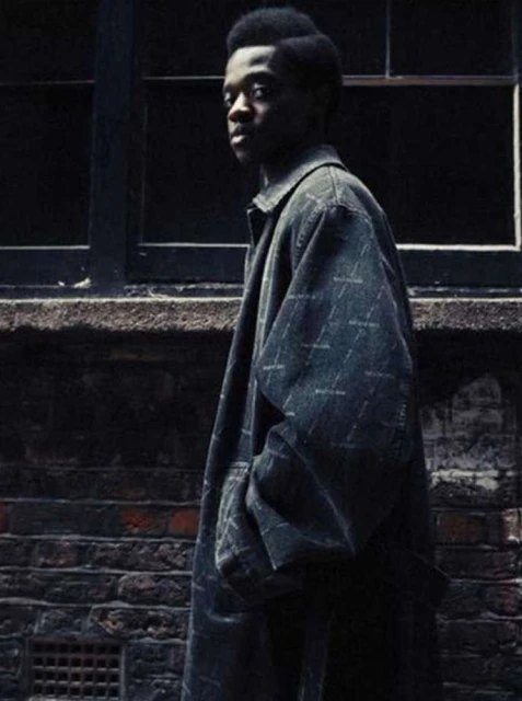 Young adult male poses in a printed overcoat in front of a brick wall and dark window, capturing urban fashion and style.