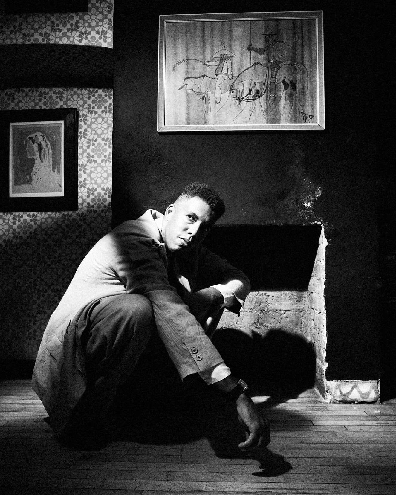 Monochrome portrait of a man squatting indoors against a dark wall with framed pictures and patterned wallpaper.