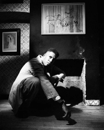 Monochrome portrait of a man squatting indoors against a dark wall with framed pictures and patterned wallpaper.