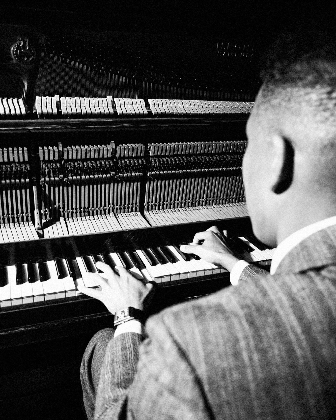 Back view of a dark-haired man in a suit playing a piano, showcasing the instrument's internal mechanics in a black and white image.