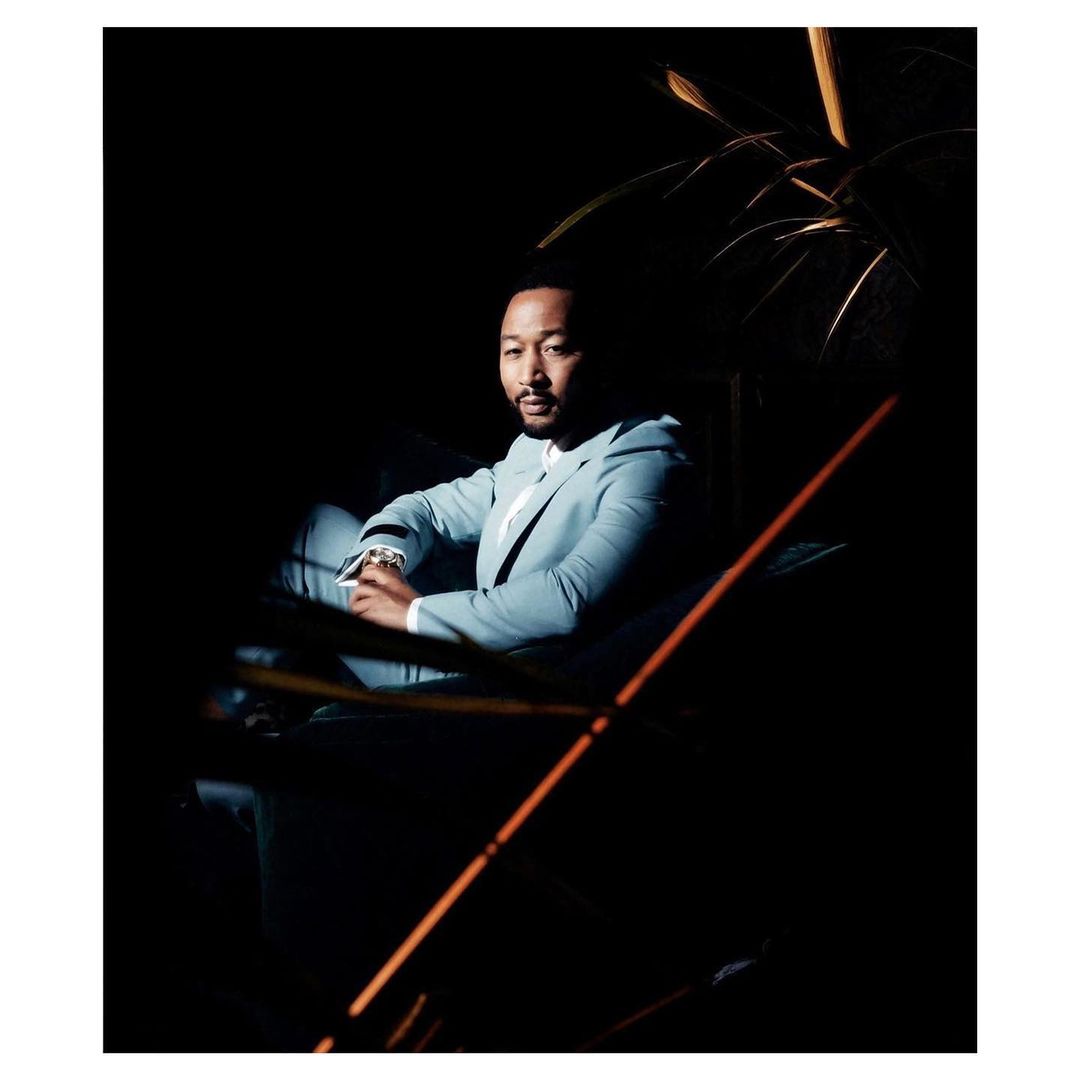 Portrait of John Legend sitting indoors, wearing a light blue suit and white shirt, captured in a dimly lit, elegant setting.