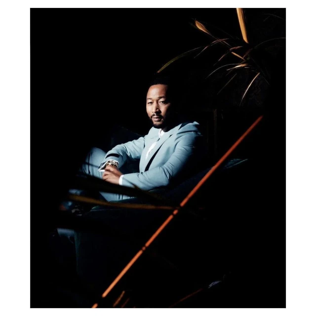 Portrait of John Legend sitting indoors, wearing a light blue suit and white shirt, captured in a dimly lit, elegant setting.
