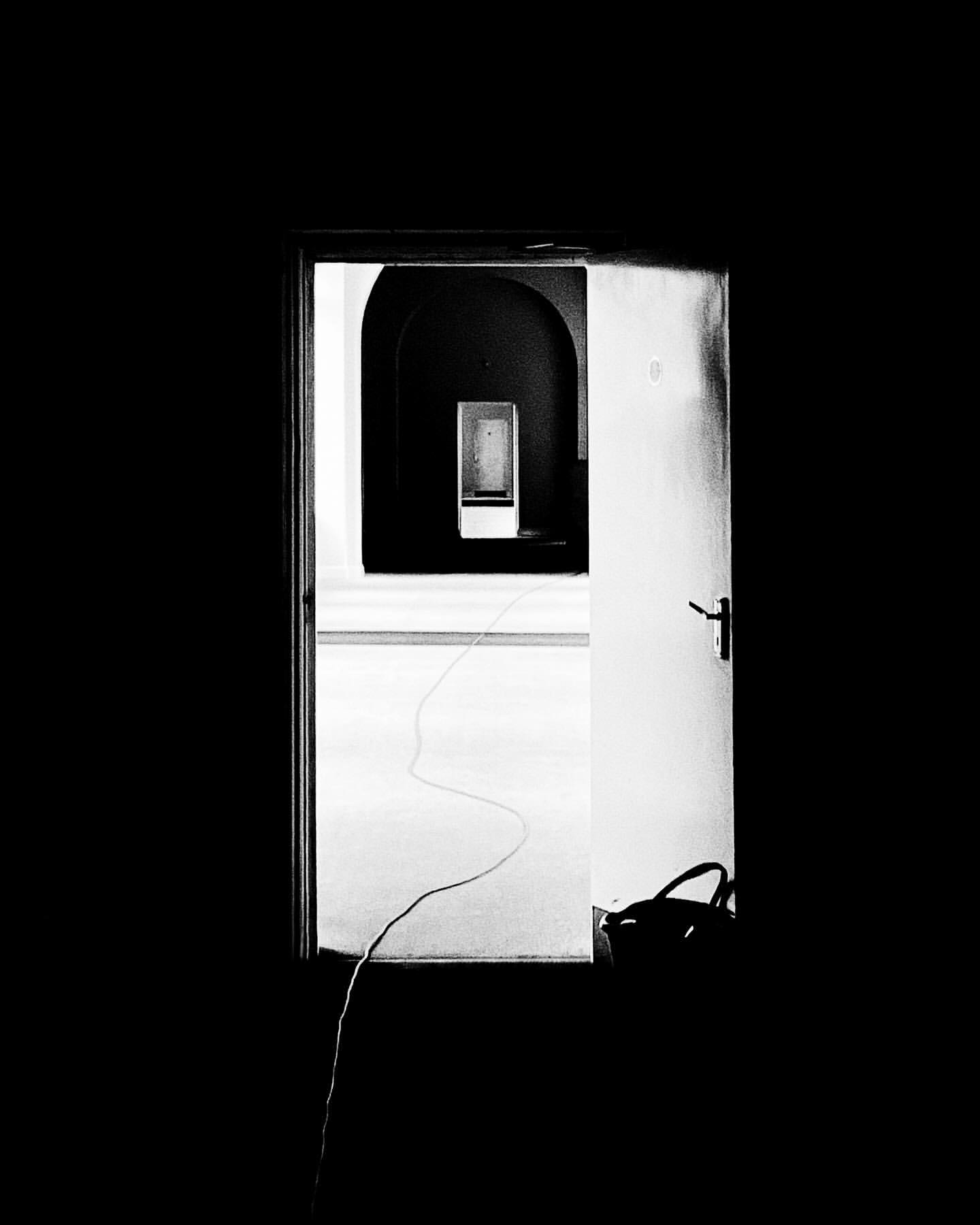 Black and white image through a doorway into a room with arched openings and a box on a shelf in the distance, high contrast.