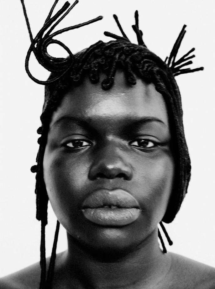 Close-up portrait of an adult female with dark skin, featuring a hairstyle of braids and strands, against a stark white backdrop.