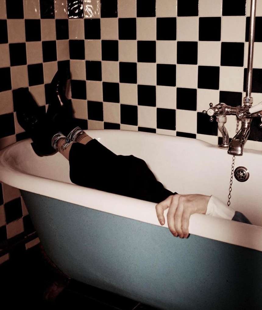 Person relaxing in a blue and white bathtub, legs up, wearing black pants, set against a checkered black and white tile wall.