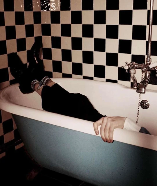 Person relaxing in a blue and white bathtub, legs up, wearing black pants, set against a checkered black and white tile wall.