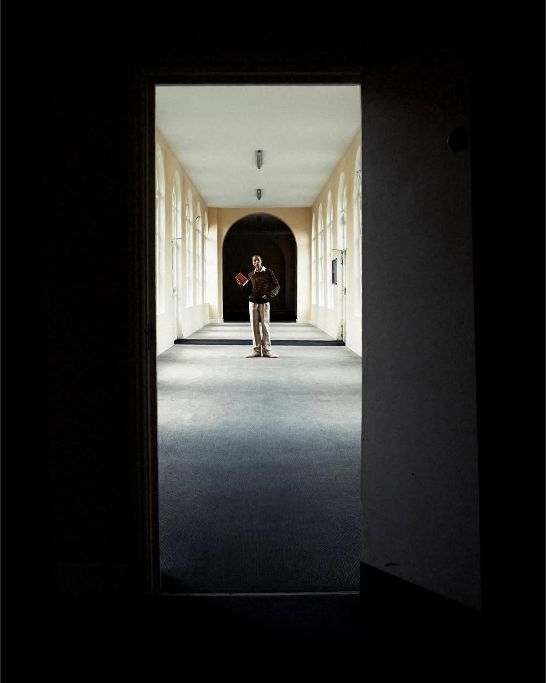 A person stands in a long hallway with archways and windows. The interior has a vintage feel with a dark gray carpet and cream-colored walls.