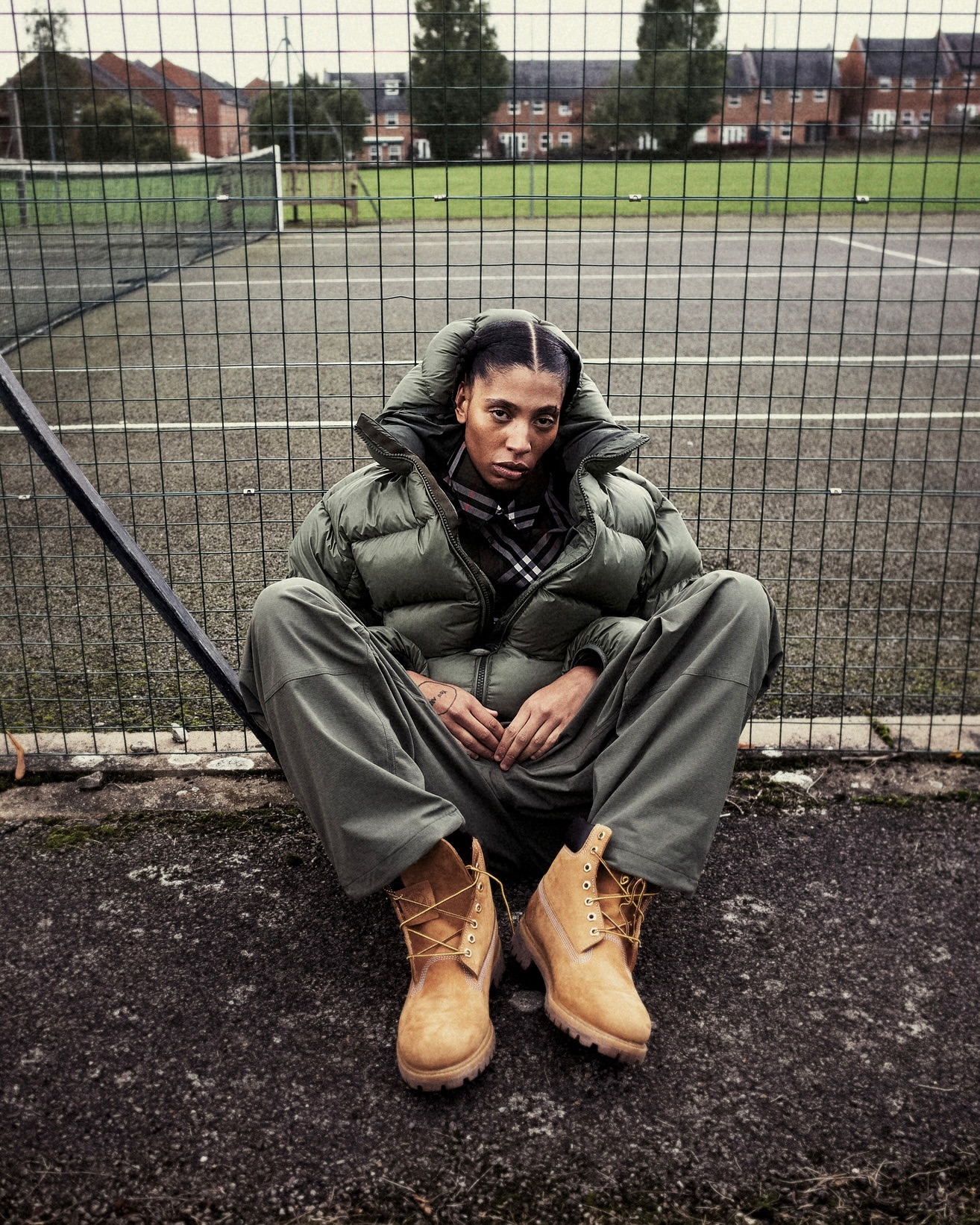 A woman in a puffer jacket and work boots sits cross-legged on a tennis court in front of a chain-link fence and buildings on an overcast day.