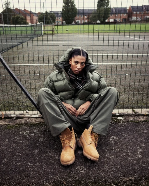 A woman in a puffer jacket and work boots sits cross-legged on a tennis court in front of a chain-link fence and buildings on an overcast day.