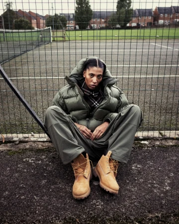 A woman in a puffer jacket and work boots sits cross-legged on a tennis court in front of a chain-link fence and buildings on an overcast day.