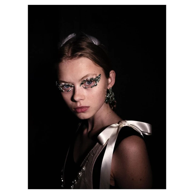 Close-up studio portrait of a young adult female model with glitter eye makeup, dark hair, and pale skin, looking at the camera.