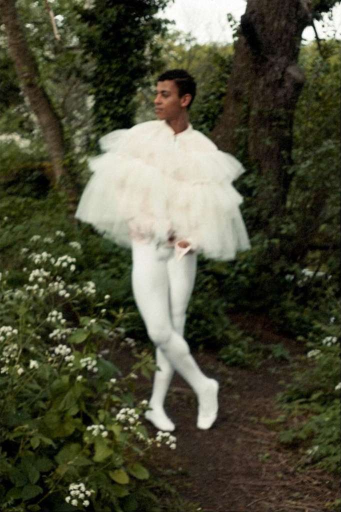 Male ballet dancer in white tulle costume posing on a dirt path outdoors in a garden with green foliage and white flowers.