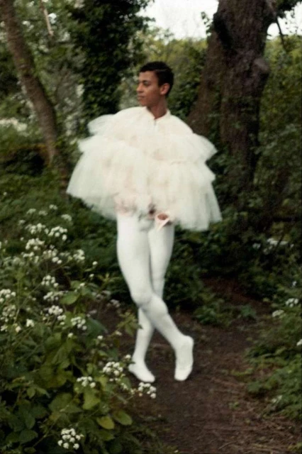 Male ballet dancer in white tulle costume posing on a dirt path outdoors in a garden with green foliage and white flowers.