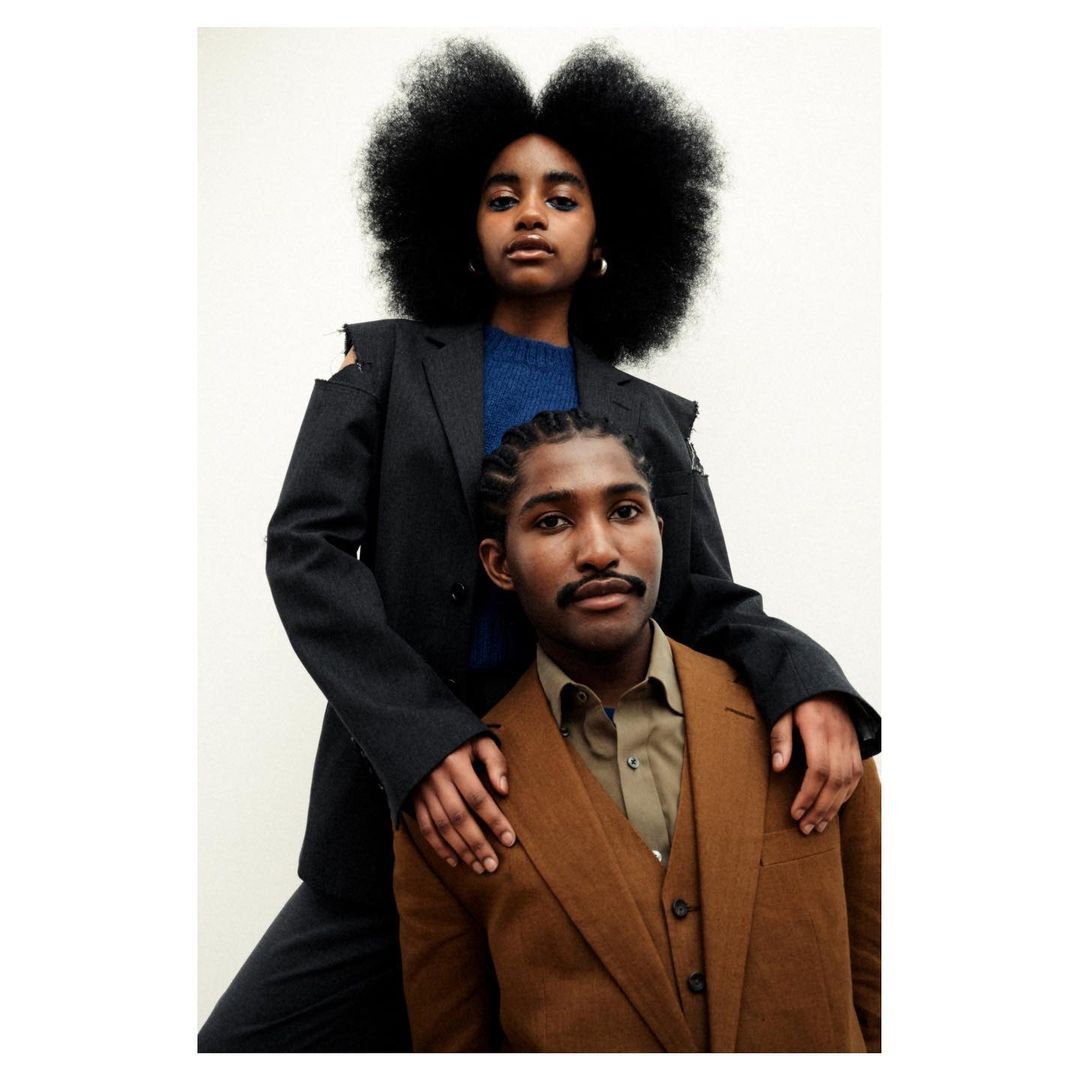 Portrait of an adult Black woman and man in suits posing indoors. The woman has large curly hair and her hand on the man's shoulder.