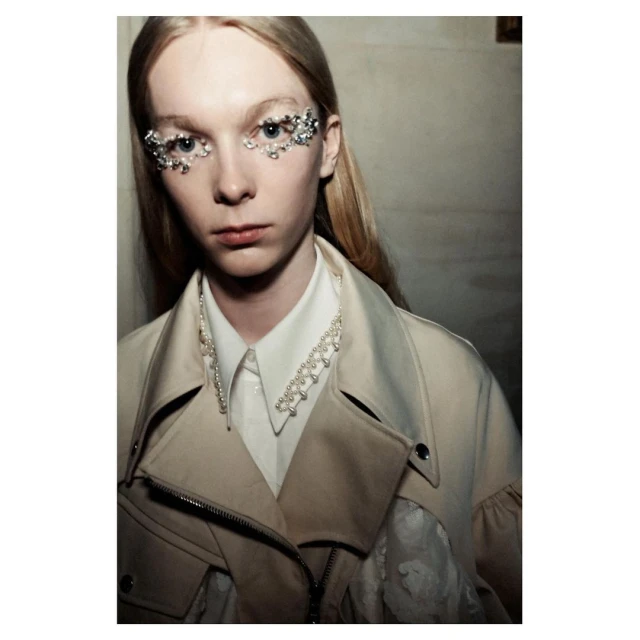 Close-up fashion portrait of a blonde adult female model with decorative jewels around eyes, wearing a white collared shirt and tan jacket.