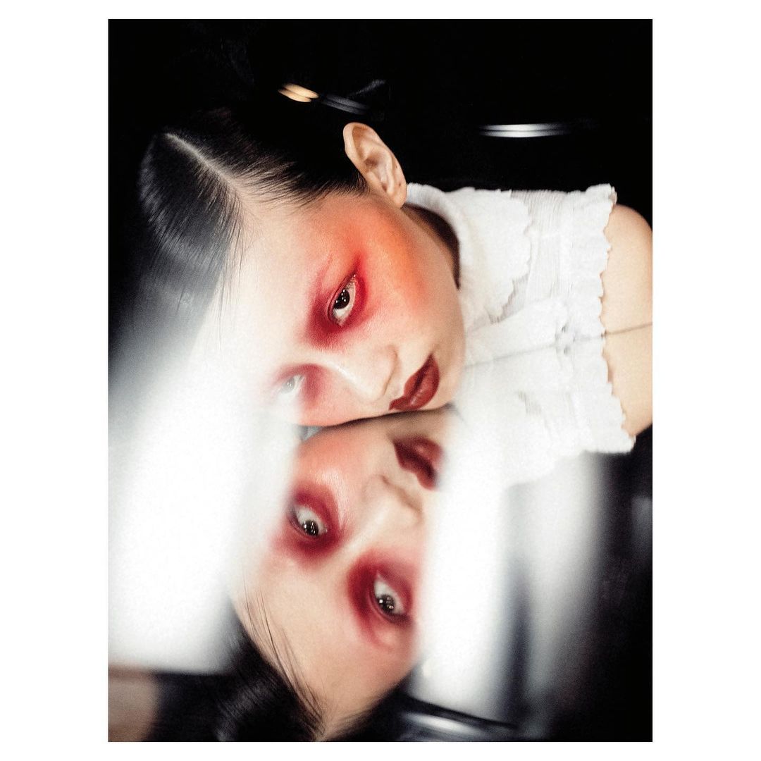 Headshot of an Asian woman with dramatic red makeup, positioned above her own reflection against a dark backdrop in a studio setting.