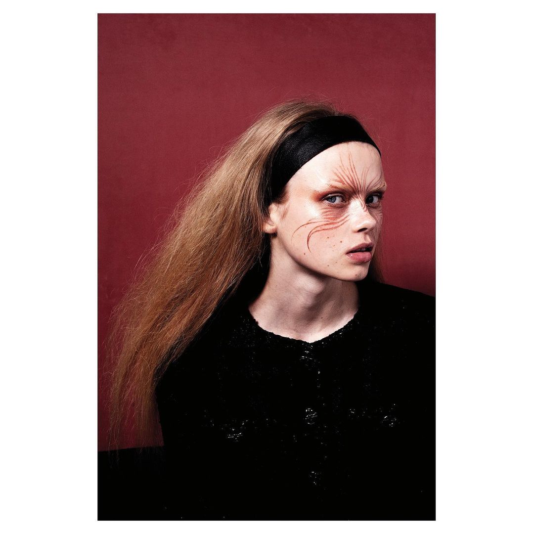 Portrait of a fair-skinned young adult woman with long brown hair, wearing a black headband and a black top, against a red backdrop.
