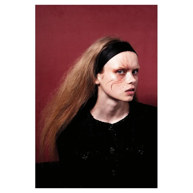 Portrait of a fair-skinned young adult woman with long brown hair, wearing a black headband and a black top, against a red backdrop.