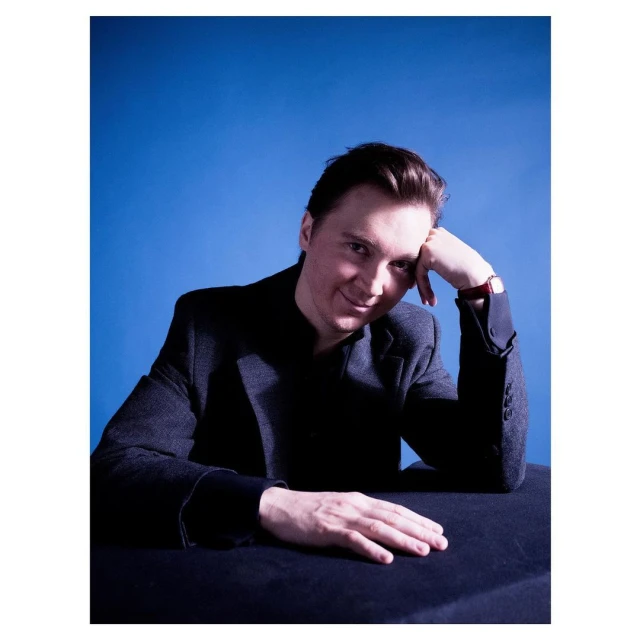 Portrait of a fair-skinned adult male wearing a dark suit and red watch, resting his head on his hand against a blue backdrop.