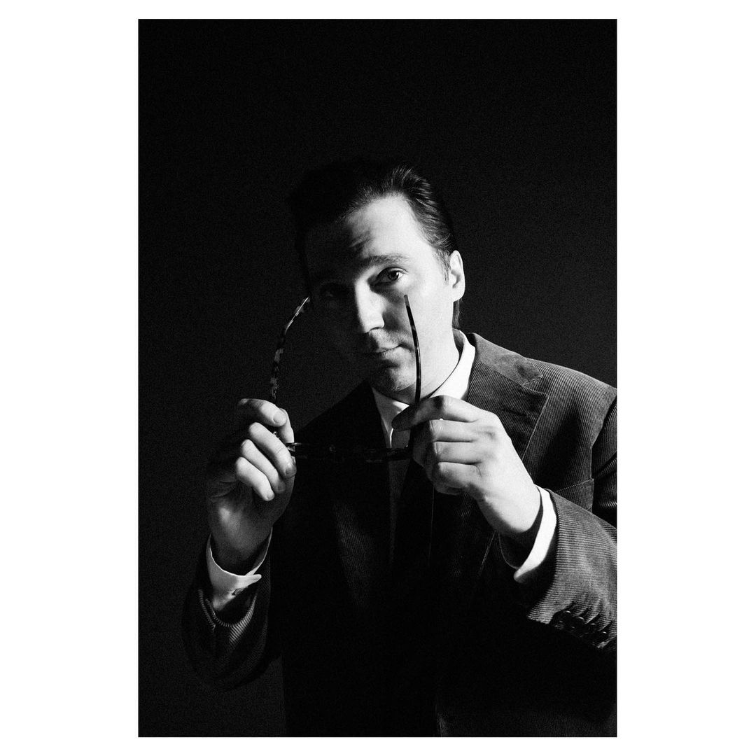 Portrait of a man in a dark suit holding glasses, photographed by Chieska Fortune Smith in black and white.