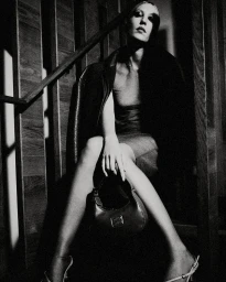 Monochrome portrait of a seated woman wearing a dark coat and holding a handbag on a wooden staircase by Chieska Fortune Smith