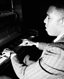 Profile portrait of a Black man playing an upright piano wearing a patterned suit jacket, photographed by Chieska Fortune Smith