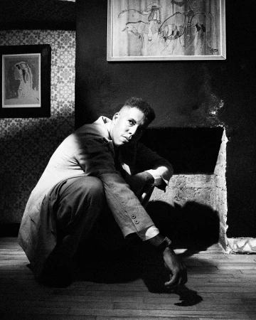 Black and white portrait of a man squatting indoors against a dark wall with framed artwork by Chieska Fortune Smith