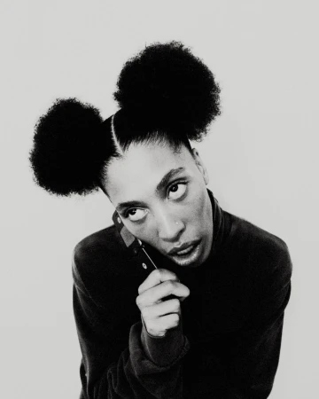 Black and white portrait of a woman with double afro puffs wearing a dark turtleneck holding a phone.