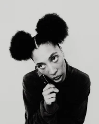 Black and white portrait of a woman with double afro puffs wearing a dark turtleneck holding a phone.