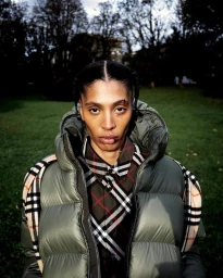Portrait of a person wearing an olive green puffer vest, plaid shirt, and draped scarf outdoors in a park.