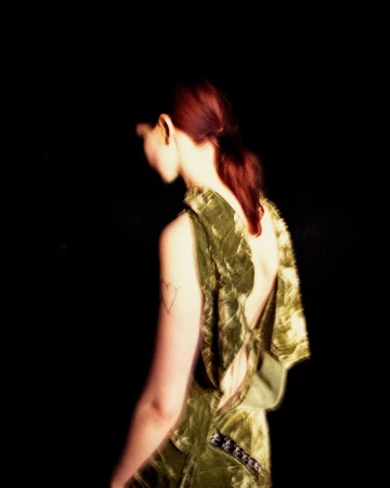 Woman with reddish-brown hair seen from behind wearing an olive green textured dress against a dark background