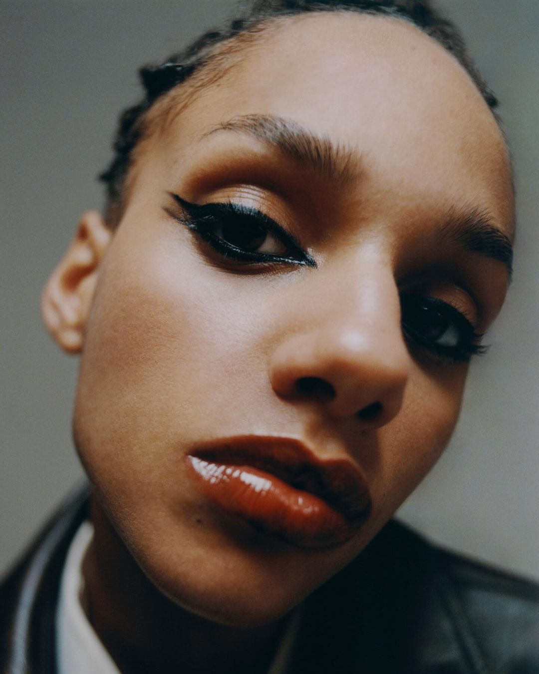 Close-up studio portrait of a young adult woman with dramatic black eyeliner and glossy red lips, wearing a black jacket.