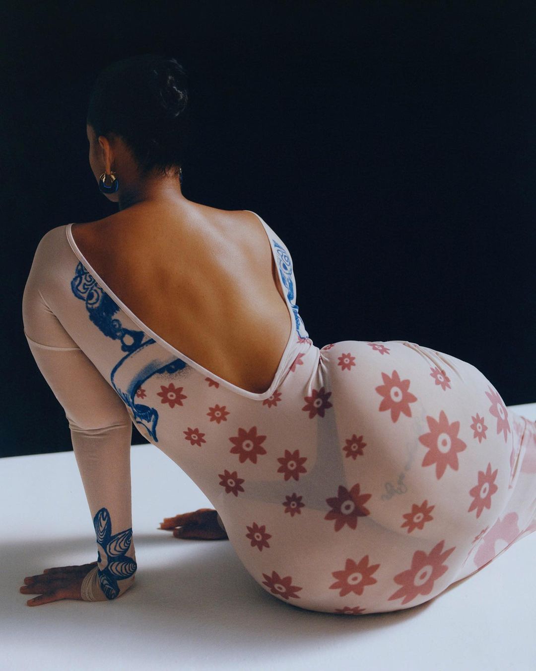 Back view of an adult female with hair up, wearing a long-sleeved, floral-patterned dress, kneeling on a white surface in front of a black background.