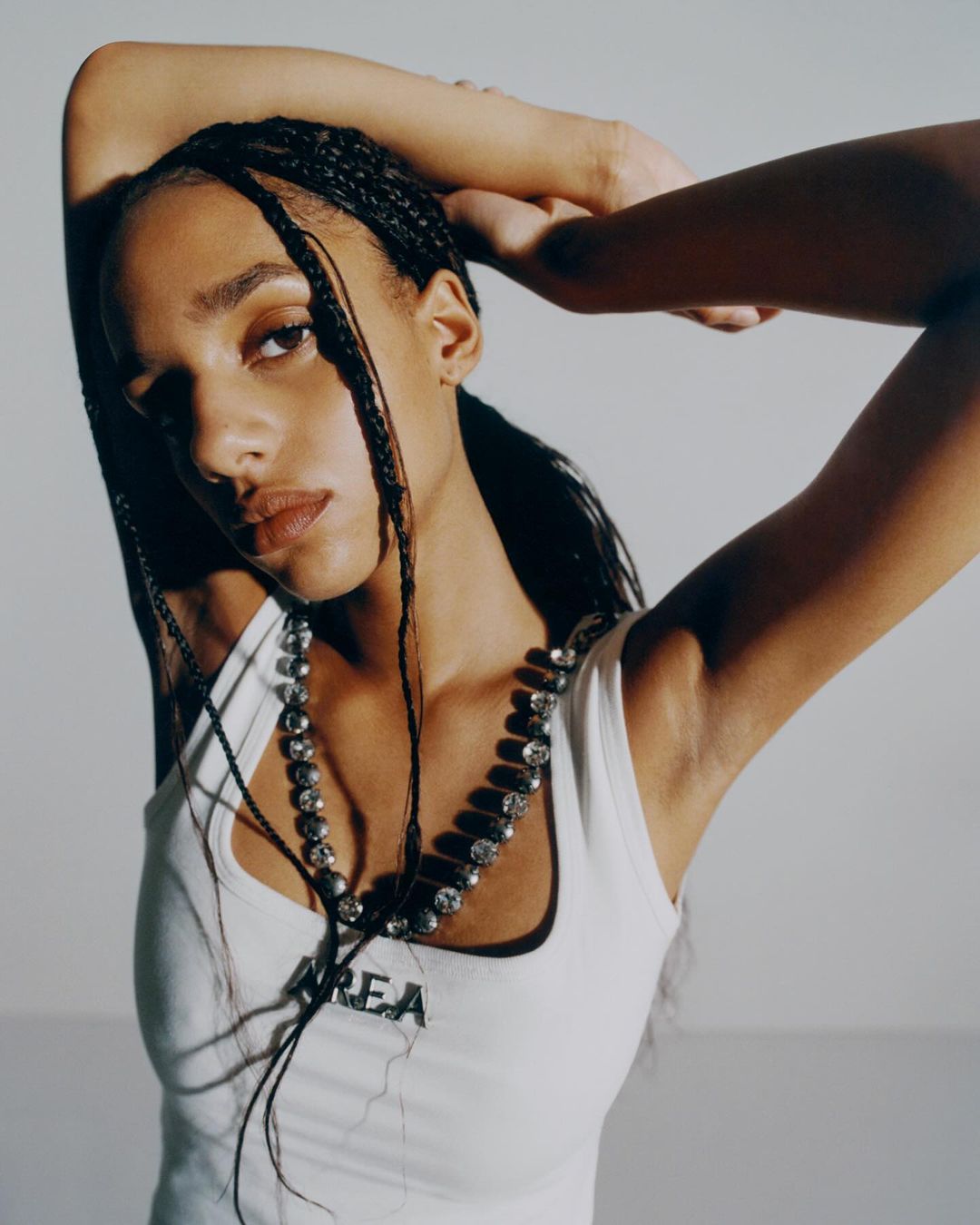 Portrait of a young woman with braided hair wearing a white tank top and beaded necklace, set against a light background in a studio.