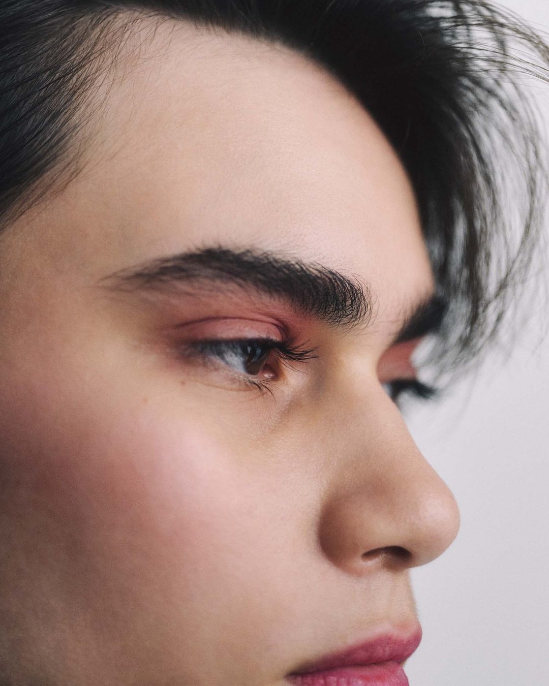 Close-up portrait of a young adult with dark hair and fair skin, featuring soft pink makeup on the eyelids and lips against a white background.