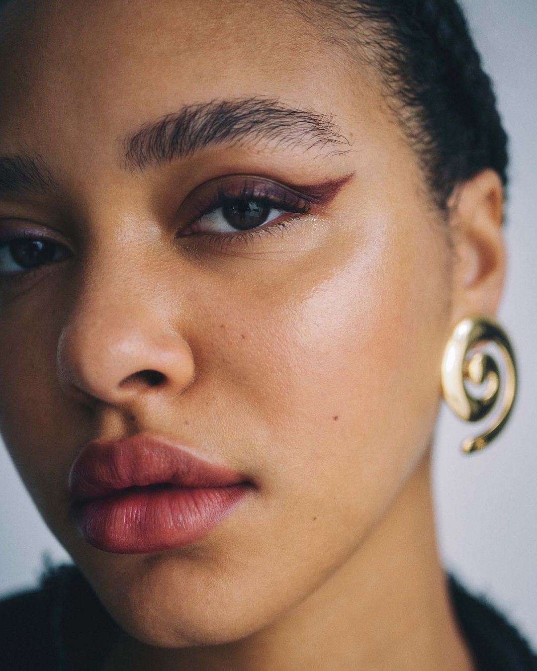 Close-up portrait of a woman with brown eyes and burgundy eyeliner, featuring gold spiral earrings against a neutral backdrop.