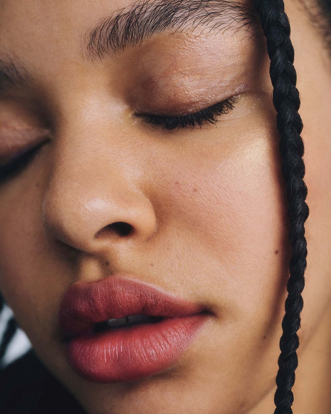 Close-up portrait of an adult woman with dark braids and eyes closed, wearing lipstick. Soft, diffused light highlights skin texture.