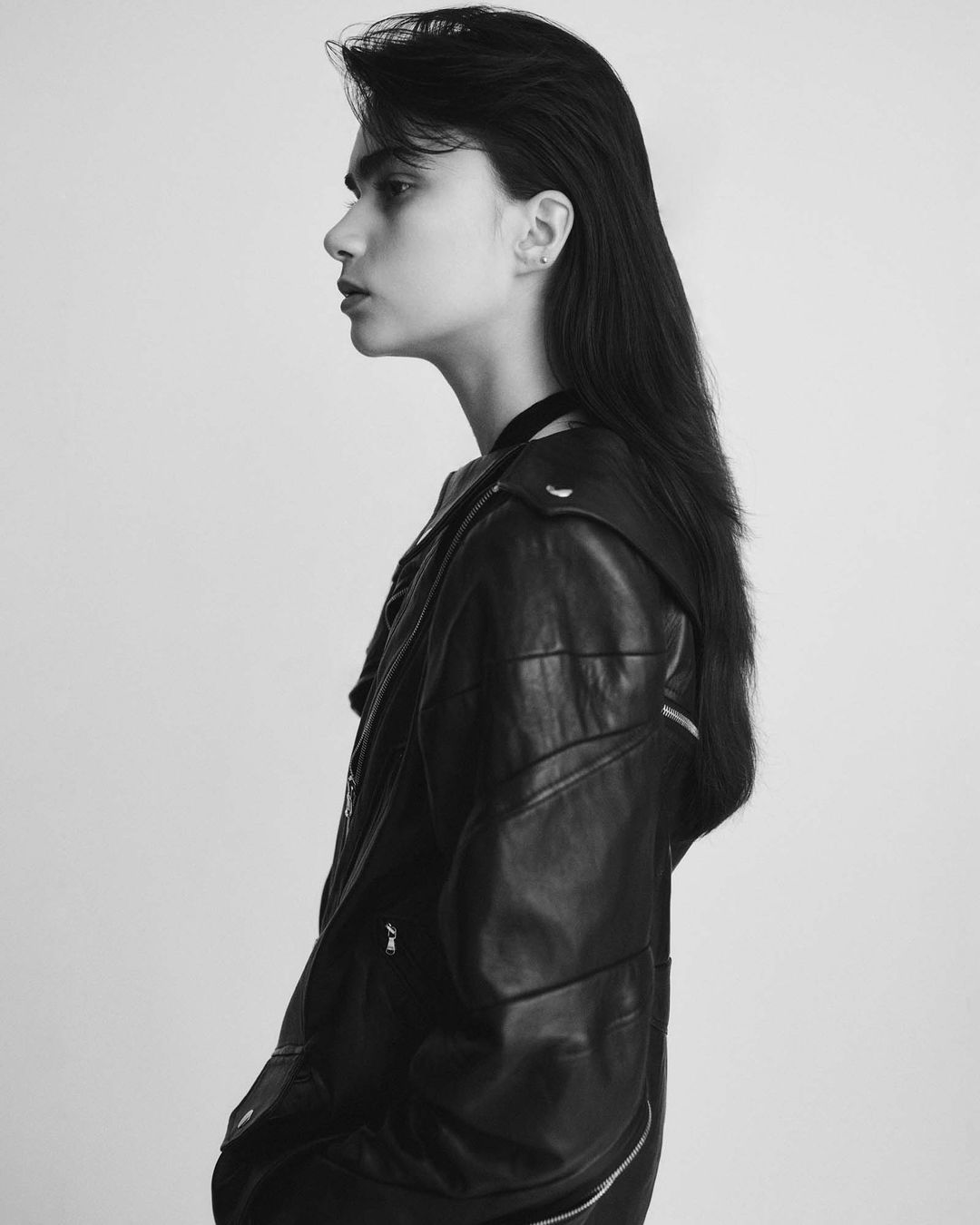 Profile shot of a young adult woman with long dark hair, wearing a black leather jacket against a light gray studio backdrop.