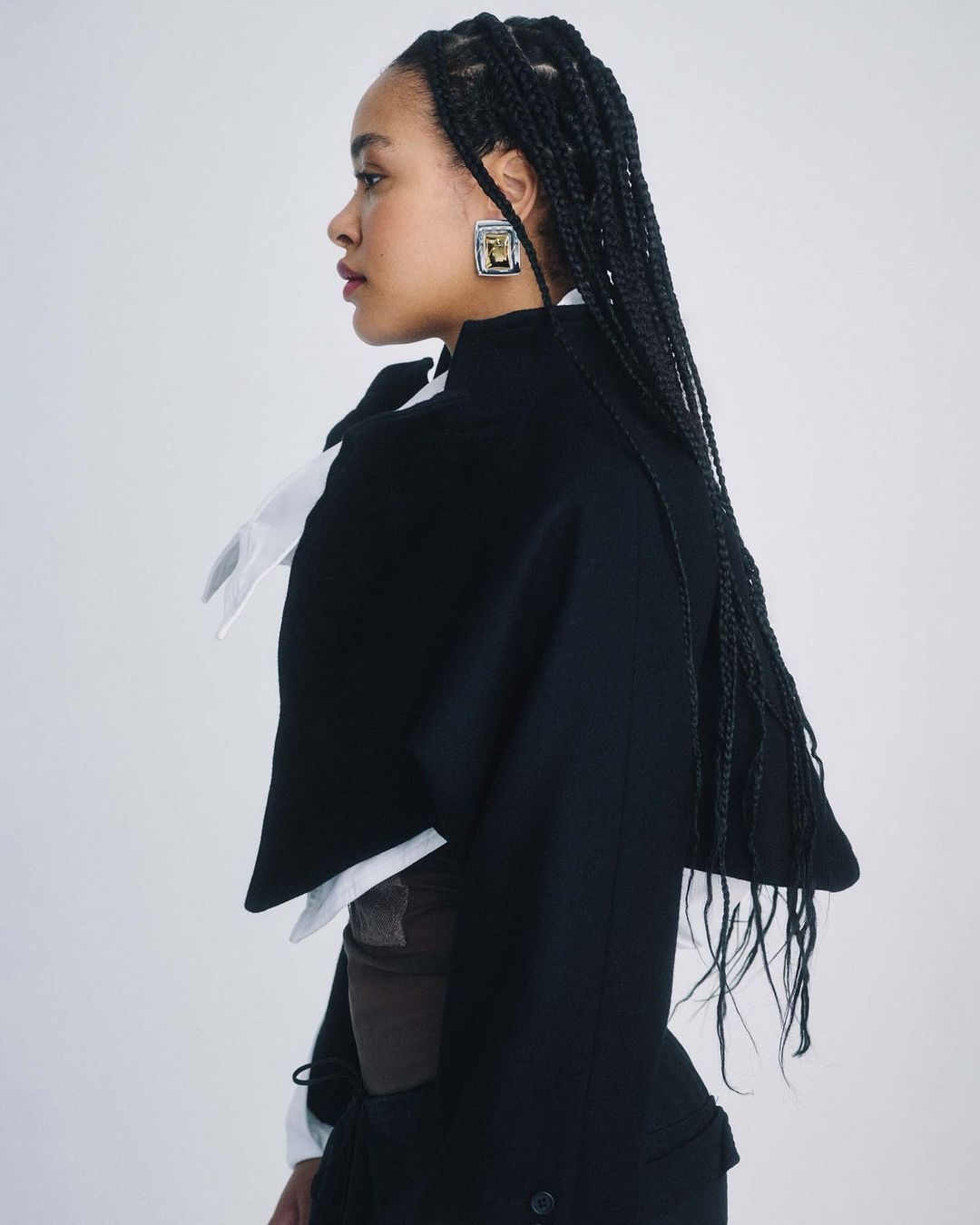 Portrait of a stylish adult woman in a black cape, white collared shirt, and statement earrings, captured in a photo studio setting.