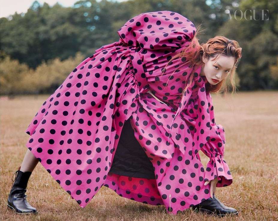 Model in voluminous pink polka dot dress and black boots bending forward on a grassy field by photographer Drew Jarrett