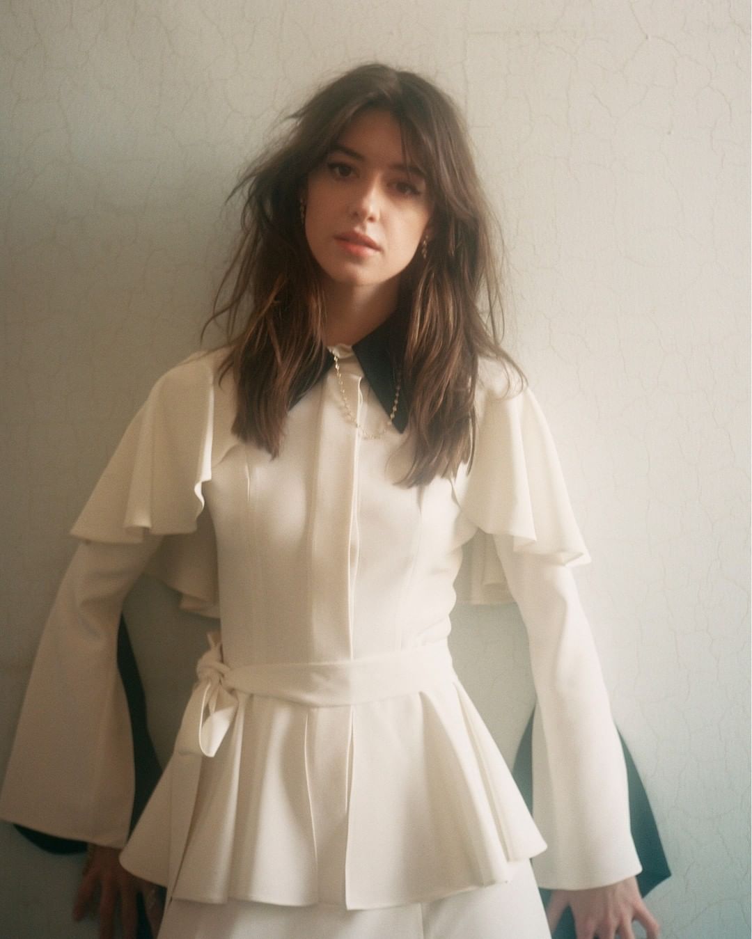 Portrait of a young woman with long brown hair wearing a white ruffled top with a black collar in front of an off-white wall.